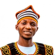 Portrait of a smiling man wearing a traditional striped cap and embroidered vest against a white background.