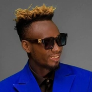 Portrait of a smiling man with blonde-tipped hair wearing sunglasses and a vibrant blue suit jacket.