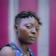 Portrait of a woman with short locs or twists looking over her shoulder against a blurred pink background.