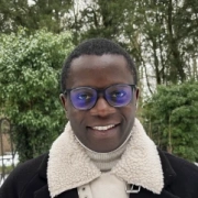 Headshot of a smiling man with glasses wearing a black jacket with a white fleece collar against green trees.